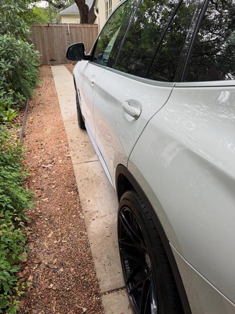 Driver side of the white BMW X3 on a narrow driveway after its hand wash, with bright paint and clean black wheels