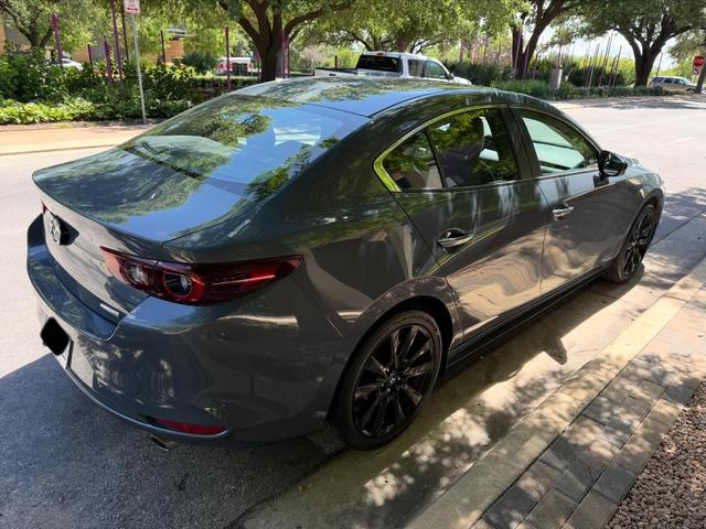 Rear three-quarter view of the gray Mazda 3 sedan in the shade, with clean taillights and glossy trunk lid